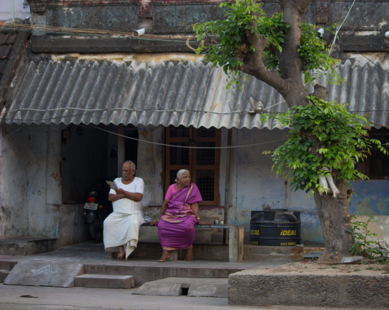 Image of an old man in a white vest and white veshti reading the newspaper. Image of old woman in a pink saree looking to her left. They sit on a bench outside a house with a tinned roof, with a tree nearby.