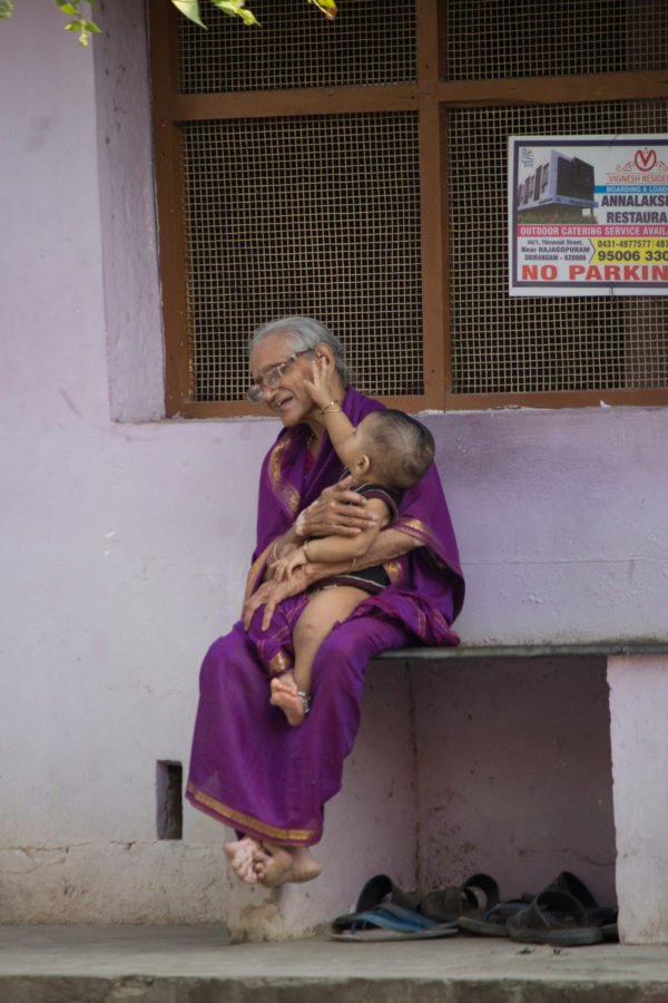 An old woman in glasses, wearing a pink saree, holds a baby that is touching her cheek. The woman has a smile on her face.