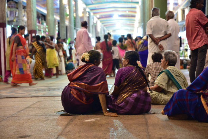 In a crowded temple hall, two women in sarees are talking to each other. 