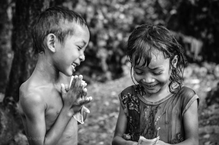 a black and white photo of two children in Cambodia, holding American dollars in their hands. They're both all smiles. One is shirtless, the other wears a dress with sequins. 